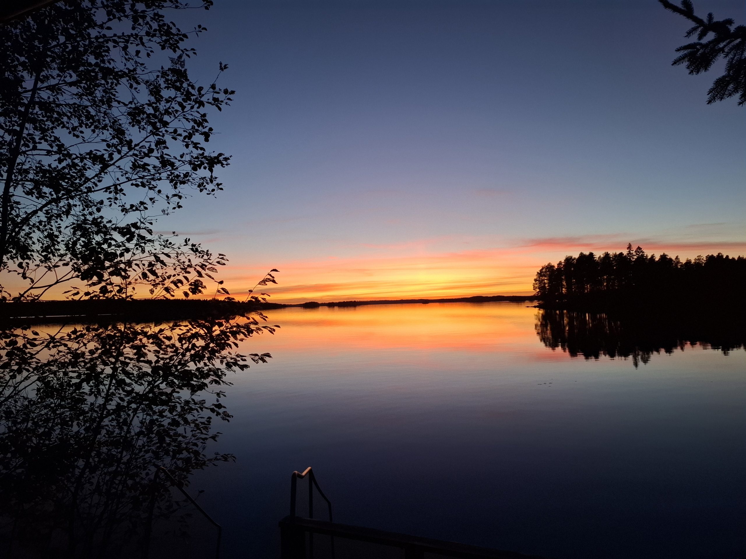 Järvimaisema laskeva ilta-aurinko. Taivaanranta kirkkaan oranssin sävyissä. Varjoihin painuneet saaret.